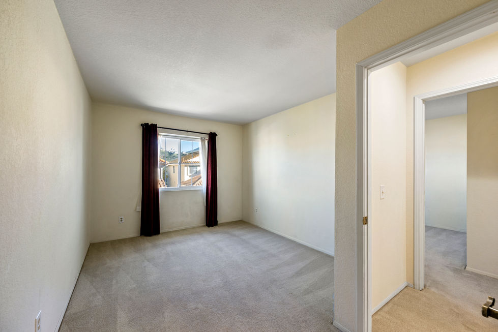 Secondary bedroom with beige carpet, neutral walls, window with dark curtains, and open doorway to hallway.
