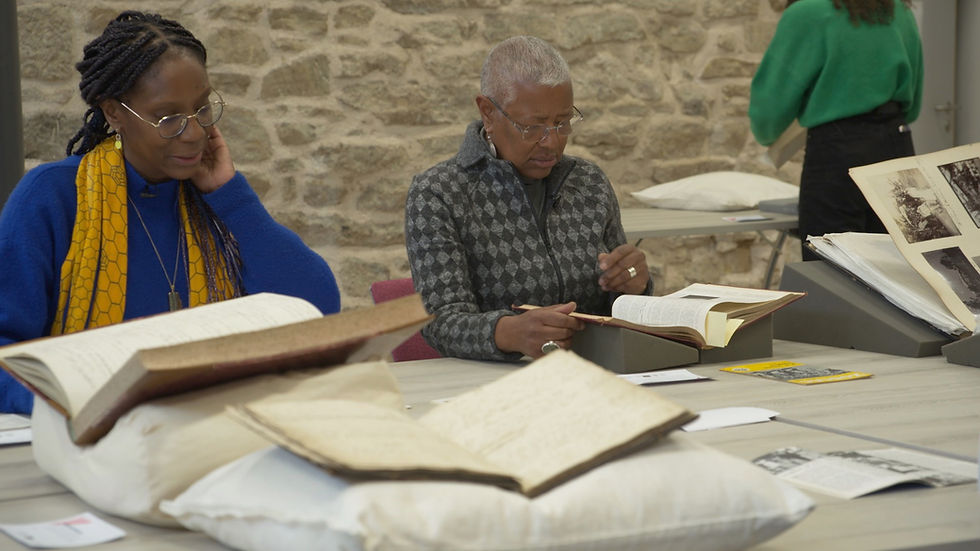 Two Black women study old books at a table in a stone-walled room, with papers and cushions around them, focused, and quiet.