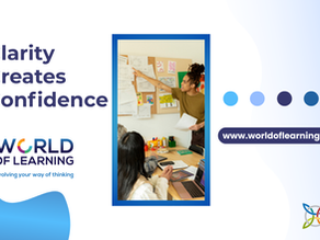 Clarity creates confidence. Photo of people sitting at a table with laptops and phones. One person is standing up and pointing to papers on a board on the wall.