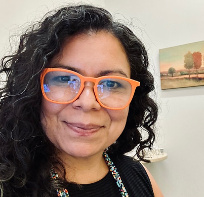 This is Carolina Rojas, she is smiling, she has curly hair and is wearing bright orange glasses and a beaded necklace, standing in a warmly lit office with artwork in the background.