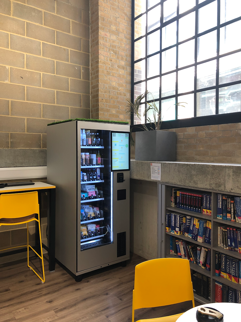 Vending machine beside a bookshelf in a modern room with brick walls and large windows. Yellow chairs add a pop of color.