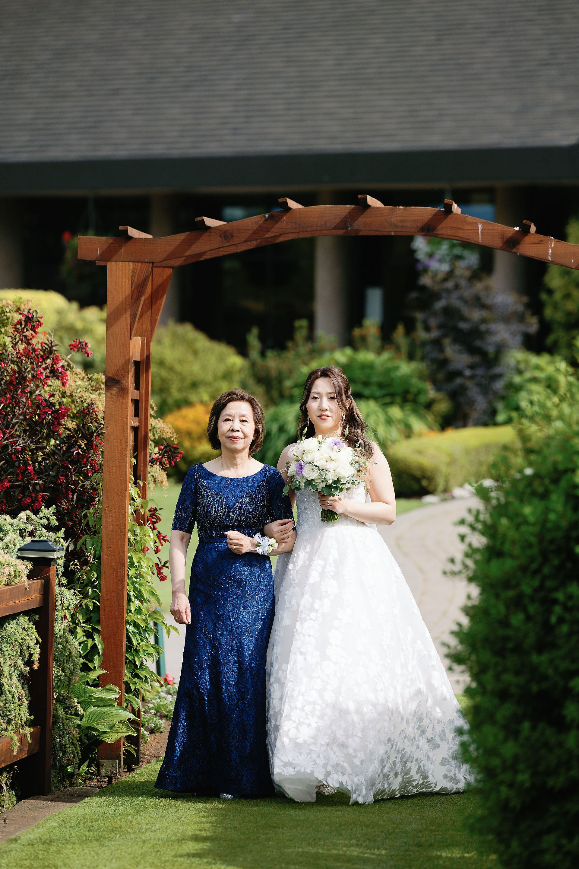 Bride walking down a garden path at Mayfair Lakes with her mother, both smiling under the wooden arbor.