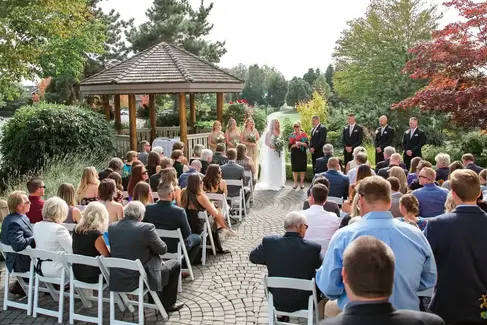 Outdoor wedding ceremony at Quilchena Golf Club with guests seated along a cobblestone aisle, facing the bride and groom under a wooden gazebo surrounded by lush gardens.