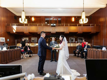 Bride and groom holding hands during their Vancouver City Hall wedding ceremony in the Council Chamber, with officiant and family guests witnessing the vows.