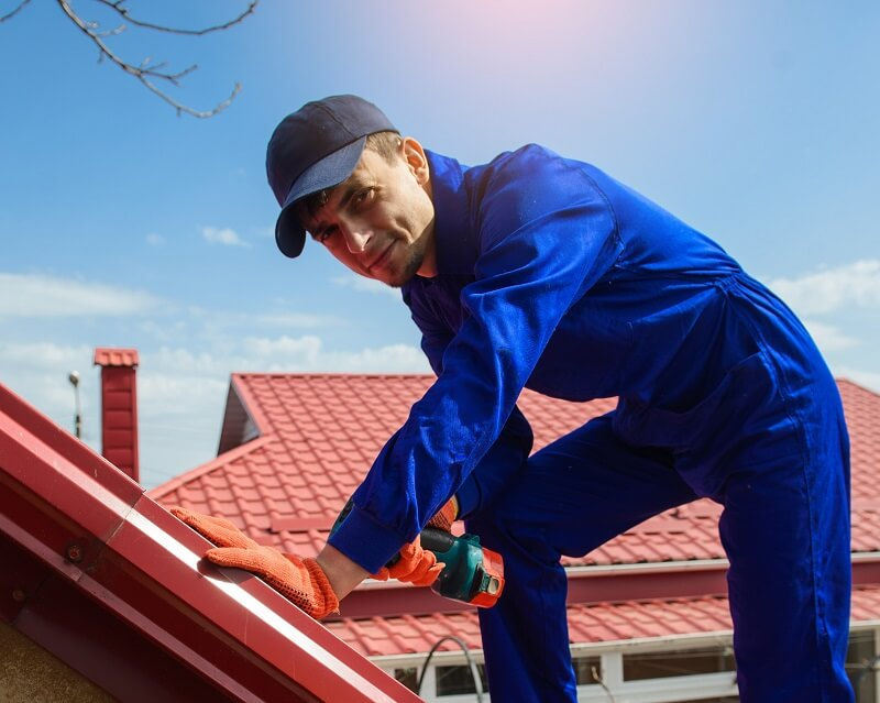 Man in blue work uniform and cap repairs a red roof with a tool. Sunny day, red rooftops in background. Focused expression.