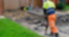 Worker in neon gear uses jackhammer on a cracked driveway. Green lawn and tools visible. Wooden garage doors in the background.
