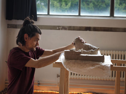 Ein mittelalter Mann kniet vor einem Tisch und schaut konzentriert die Tonmasse an, die er formt