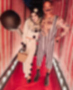 Couple in clown costumes, standing in a red-carpeted hallway with lights.