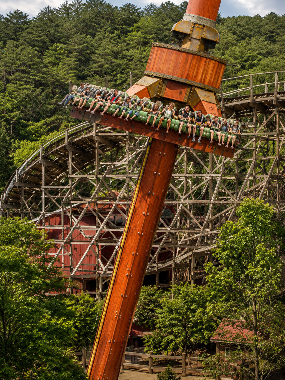 Passengers hang on during dollywood's timber tower ride shown against a forest backdrop, with a wooden roller coaster behind.