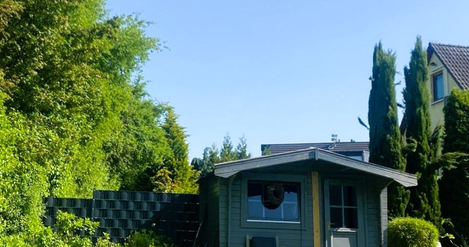 Garten mit Rasenfläche, blühenden Beeten und einem kleinen grauen Gartenhaus unter klarem, blauem Sommerhimmel. Einfamilienhaus Langenfeld