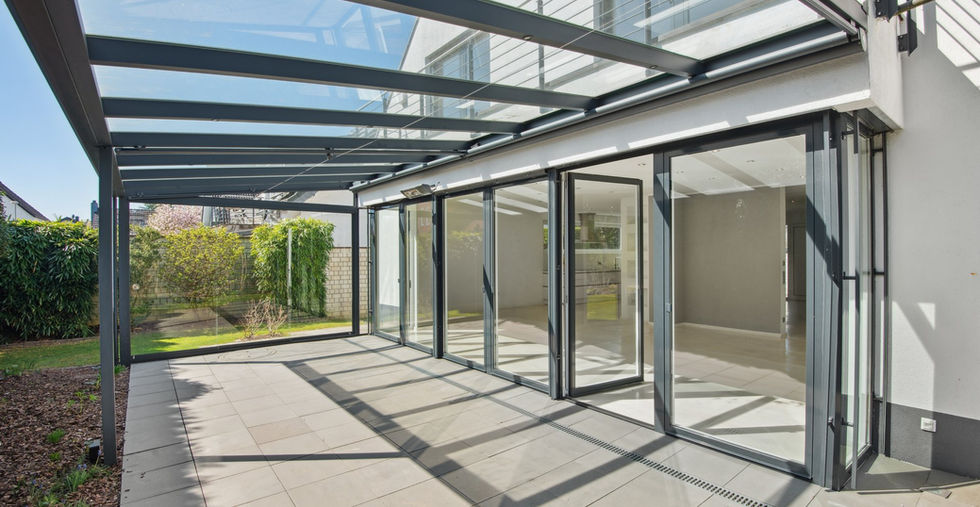 Großzügige, überdachte Terrasse mit moderner Glasdach-Konstruktion und integrierten LED-Spots in den Querstreben; bodentiefe Flügeltüren führen direkt in den lichtdurchfluteten Wohnbereich; gepflegter Garten mit Sichtschutzbepflanzung im Hintergrund.