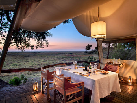 A luxurious safari tent dining setup at sunset, with a white tablecloth-covered table set for two, surrounded by wooden chairs and lanterns, overlooking a vast grassy savannah under a colorful evening sky.