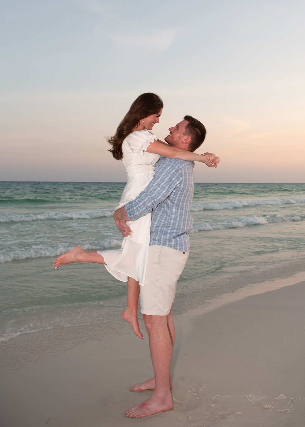 Couple in white and blue embrace on a beach at sunset, smiling as waves roll behind them.