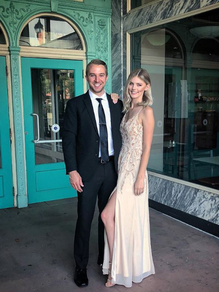 A smiling man in a suit and tie stands with a woman wearing a long gown with a slit, who is resting her hand on his shoulder.