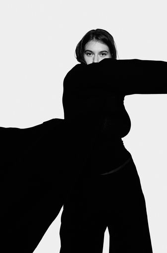 kontrastreiches Schwarzweiß Halbportrait einer jungen Frau in Silhouette mit schwingendem dunklen Mantel und ausgestrecktem Arm vor weiß