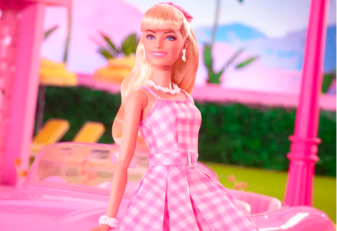 A fair skinned barbie wearing a checkered pink and white dress with pearl necklace, earrings and bracelet sat by her pink car with a background of her dreamhouse with green hedges, yellow lawn chairs.
