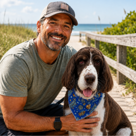 Smiling man kneels with a brown-and-white dog in a blue bandana on a sunny beach boardwalk.