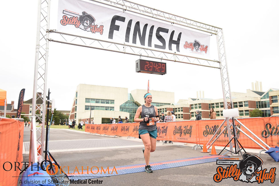 Woman crossing finish line. Above her, a banner reads "Stilly Half Finish." Time is 2:34:29,