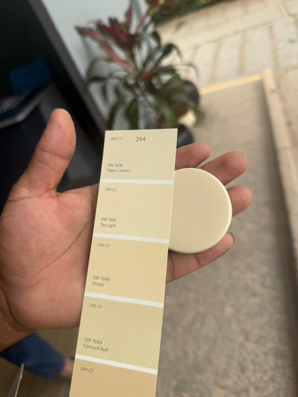 A custom tan ceramic sample held against a Sherwin Williams paint strip containing 'Totally Tan' and 'Caramelized', demonstrating how custom lighting finishes can be coordinated with neutral wall colors.