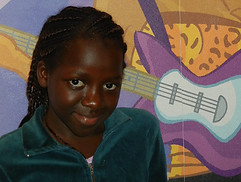 8 year old Nigerian girl sitting down, looking into the camera, with painting in the background