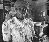 Black & white photograph of African-American woman wearing a patterned dress and headscarf, in a poorly furnished room