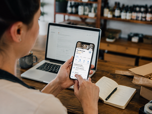 Woman at a wooden desk reviews a business profile on her phone beside a laptop, notebook, and packing supplies in a cozy cafe workspace