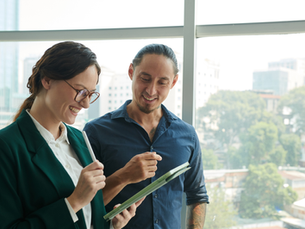 Two coworkers smile as they review a tablet with a stylus in front of a bright office window overlooking the city.