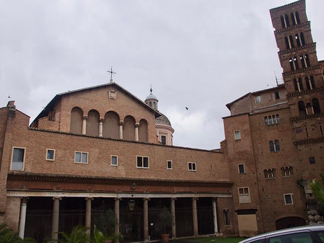 The brick exterior of the Basilica of Santi Giovanni e Paolo in Rome, showcasing a lower portico with classic columns and an upper facade with five arched niches. To the right, a multi-tiered medieval brick bell tower rises against a cloudy sky.