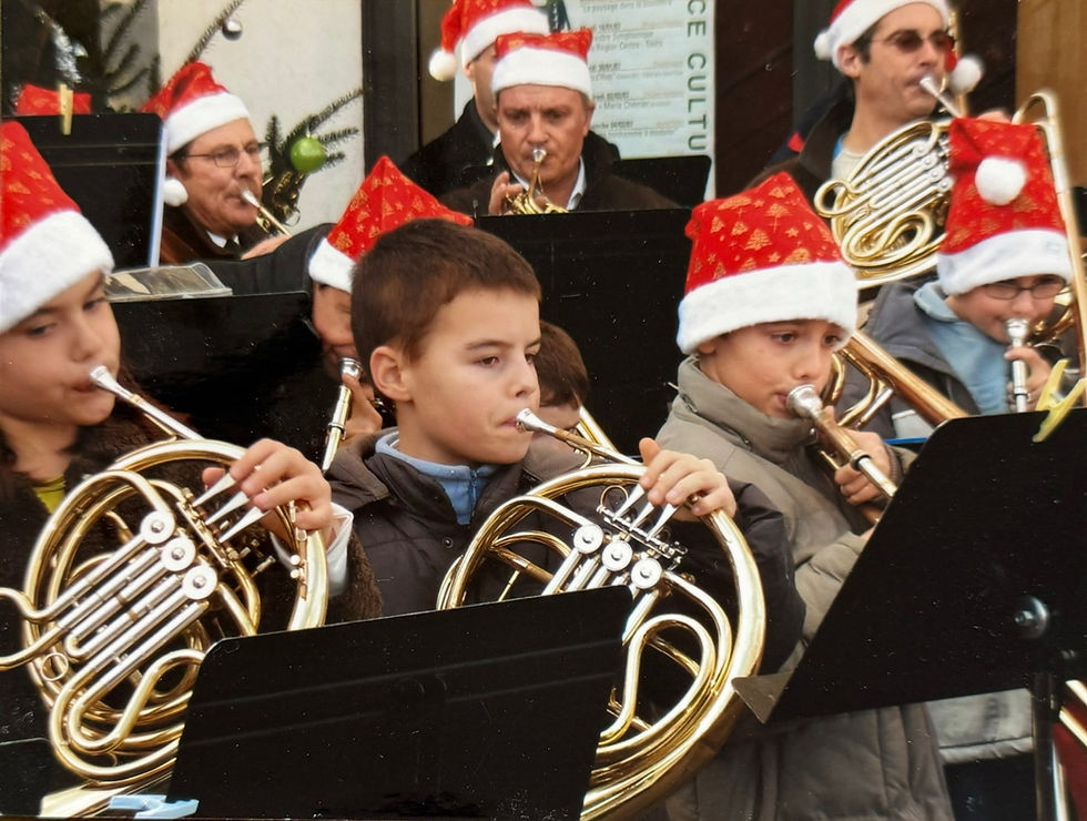 Des enfants portent des bonnets de Noël et jouent du cor et trombone lors d'une fanfare en extérieur. L'ambiance est festive et joyeuse.