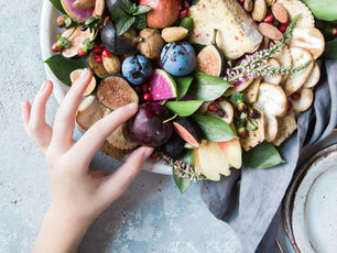Hand reaching for fruit platter with figs, grapes, pomegranate, and crackers on leafy green bed. Blue-grey cloth and plate nearby.