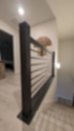 Modern indoor staircase with black metal railing, tan walls, and light wood flooring. Wall art visible above. Soft, neutral lighting.