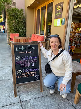 Christine Evans, author with short brown hair and sunglasses on her head, crouched by a sign at Linden Tree books