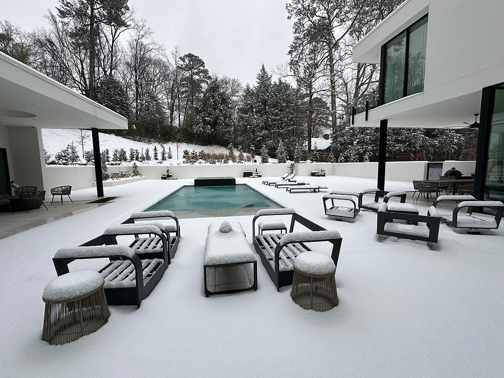 Modern patio covered in snow, featuring black furniture and a central pool. Surrounded by snowy trees under a gray sky, creating a serene mood.