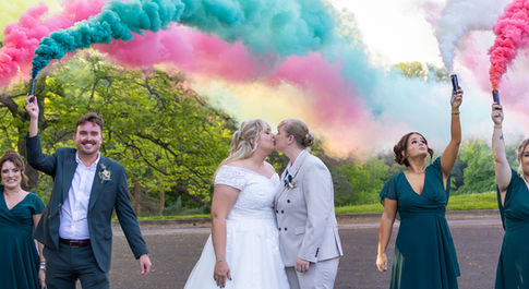 Brides kissing while friends hold colored smoke flares at Rushpool hall.
