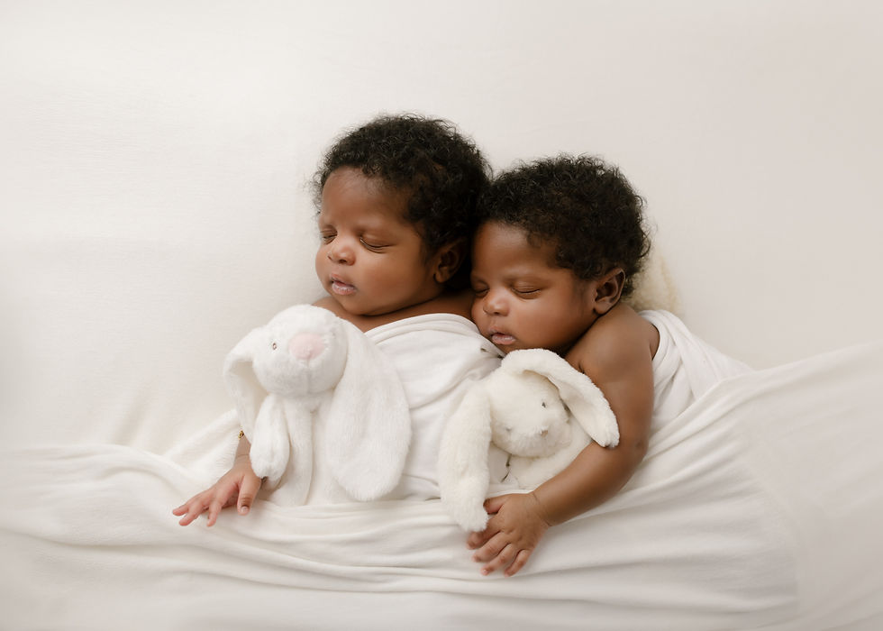 Two babies sleep peacefully, cuddling white plush bunnies, wrapped in soft white blankets on a light background.
