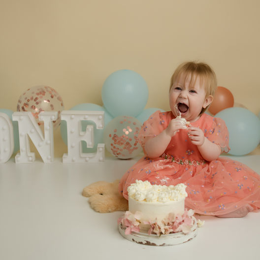A joyful celebration as a little one dives into her first birthday cake, surrounded by pastel balloons and a decorative "ONE" sign