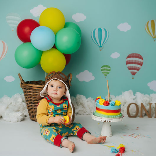 First birthday cake smash in Bristol featuring a baby in an aviator outfit sitting beside a rainbow birthday cake on a hot air balloon themed backdrop.