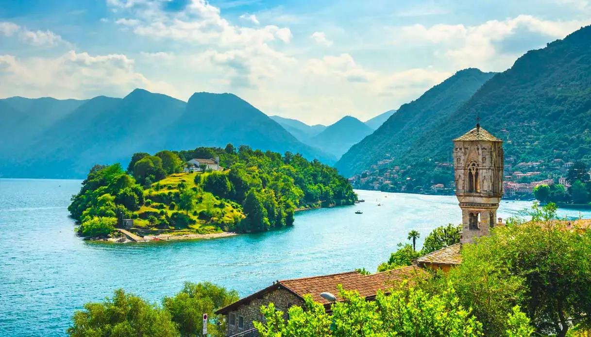in primo piano campanile di Ossuccio tra vegetazione, al centro dell'immagine lago di como con isola comacina. sullo sfondo le montagne comasche