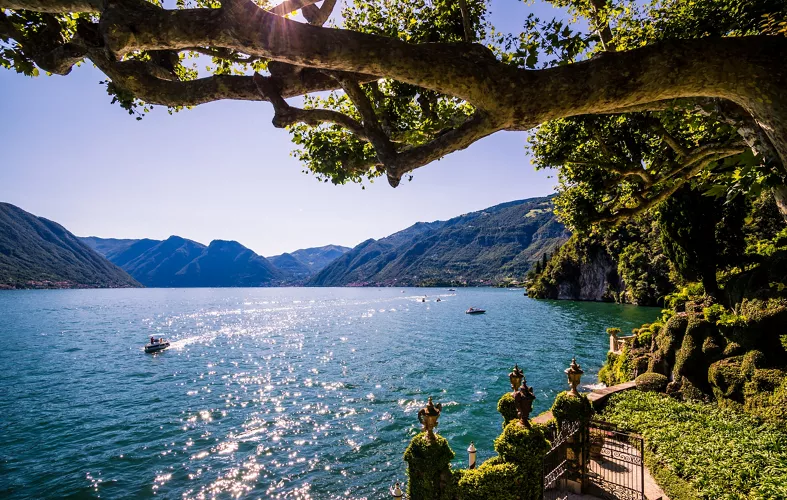 in primo piano ramo di albero con foglie verdi e giardino con cancello. sullo sfondo il lago blu con barche e montagne