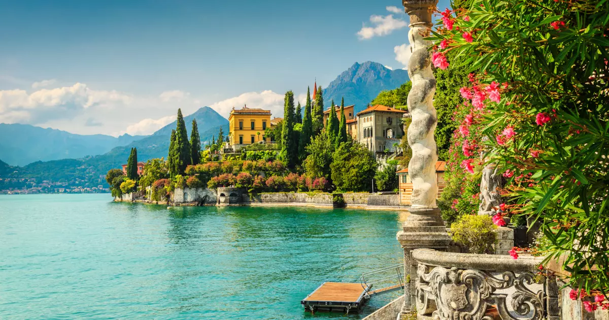 Paesaggio del Lago di Como, acqua azzurra, villa gialla con alberi verdi in una giornata soleggiata. montagne sullo sfondo con cielo blu con poche nuvole bianche. in primo piano una colonna con dei fiori rosa