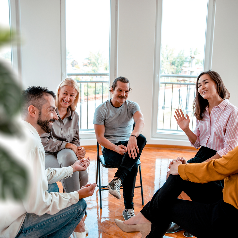 Group of five people sitting in a bright room with large windows, smiling and chatting. Casual, friendly atmosphere.