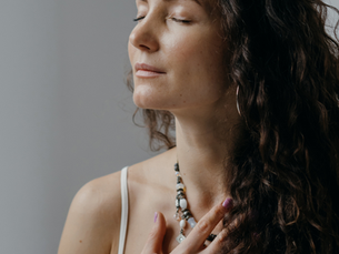 Woman with curly hair, eyes closed, wearing a white top and necklace, touching her chest. Calm expression, neutral background.