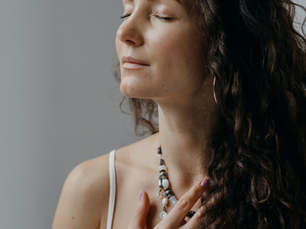 Woman with curly hair, eyes closed, wearing a white top and necklace, touching her chest. Calm expression, neutral background.