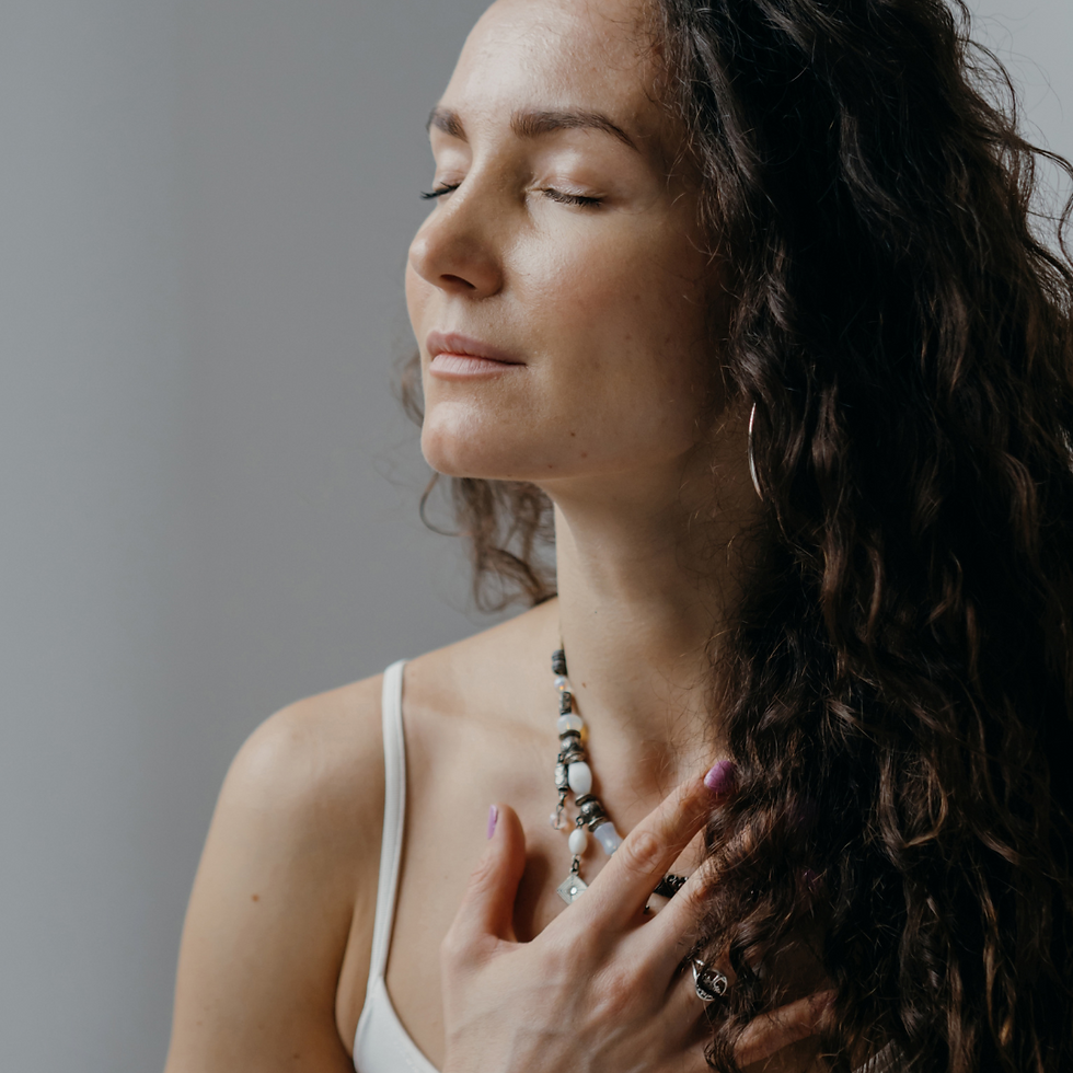 Woman with curly hair, eyes closed, wearing a white top and necklace, touching her chest. Calm expression, neutral background.