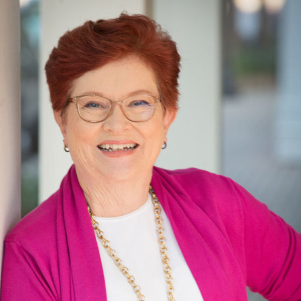 Smiling woman with red hair and glasses, wearing a pink jacket and gold necklace. Bright, blurred background suggests indoor setting.