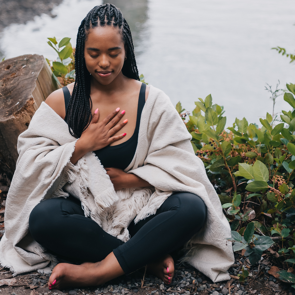 A woman meditates by a lakeside, eyes closed, hand on chest, wrapped in a beige blanket. Greenery surrounds her, conveying calmness.