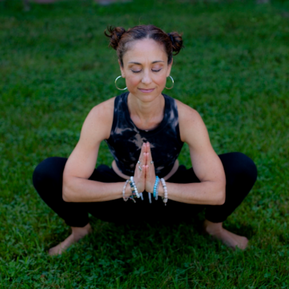 Woman squatting on grass with eyes closed, hands in prayer position. Wearing black tie-dye top, black pants, bracelets. Calm and peaceful.