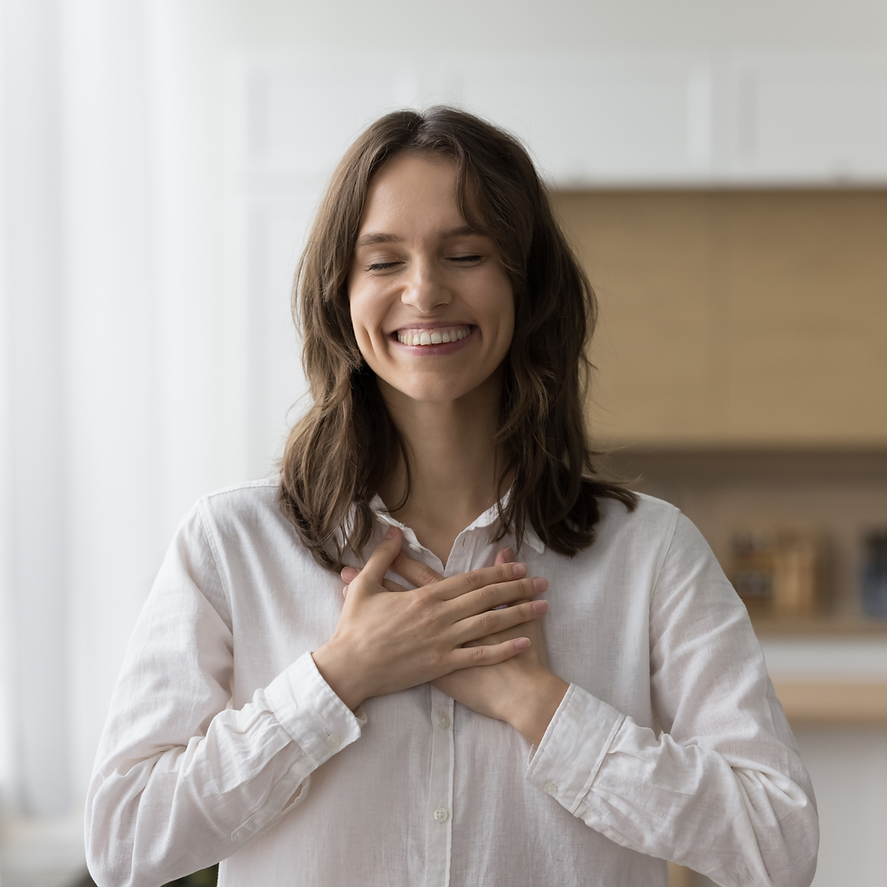 Smiling woman with closed eyes holds hands on chest, expressing gratitude. She wears a white shirt in a softly lit, blurred indoor setting.