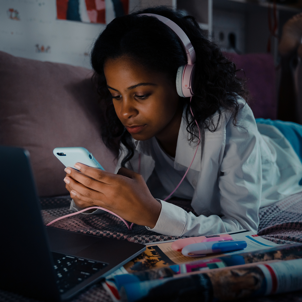 Young woman with headphones focused on smartphone, lying on bed. Nearby are an open laptop, magazines, and colorful markers. Dimly lit room.
