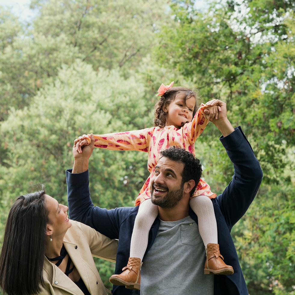 Man holding a smiling child on shoulders, woman reaching out to child in a park. Child in pink dress, trees in the background, joyful scene.